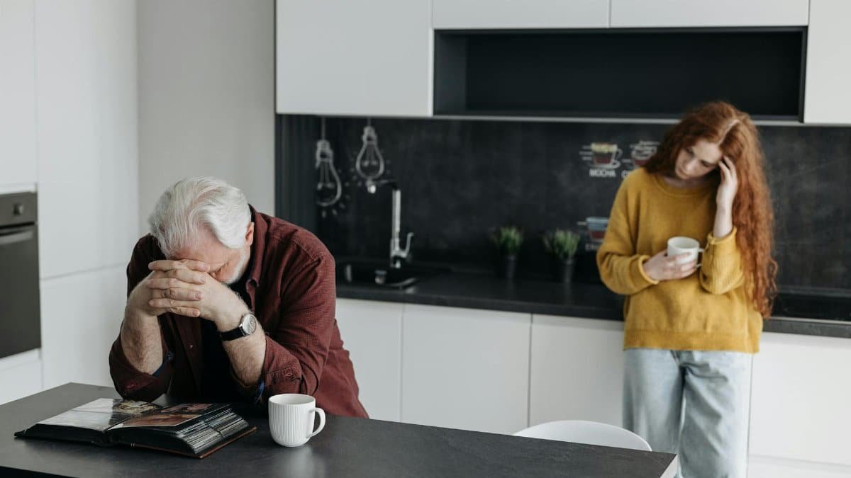 A man grieving at a kitchen table with a woman nearby, conveying emotions of loss and reflection.