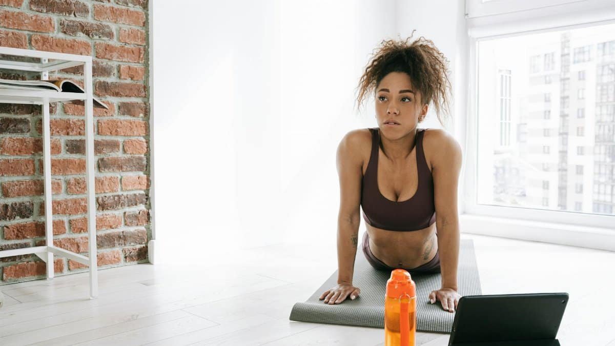 Woman in activewear doing yoga indoors with tablet and water bottle nearby.
