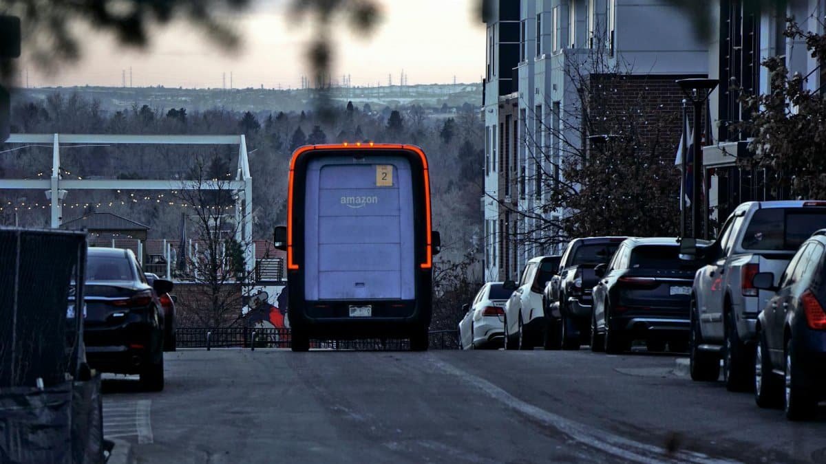 Amazon delivery van driving through an urban street at twilight with cars parked along the side.