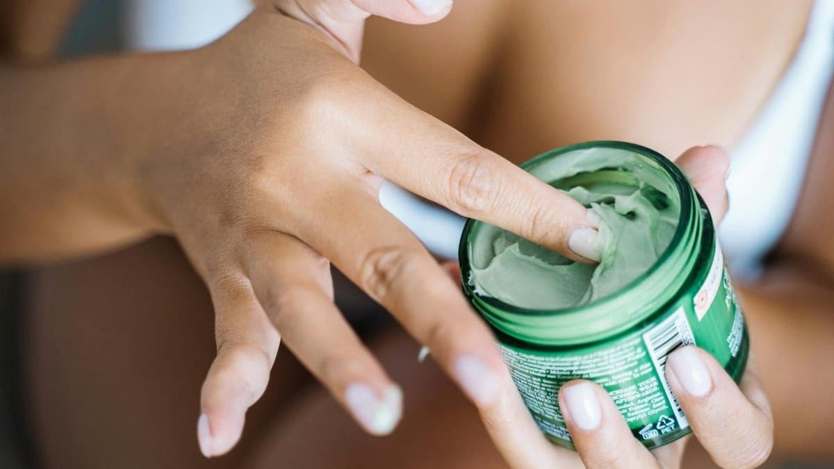 Close-up of a woman's hand applying cream from a green jar, focusing on skincare routine.