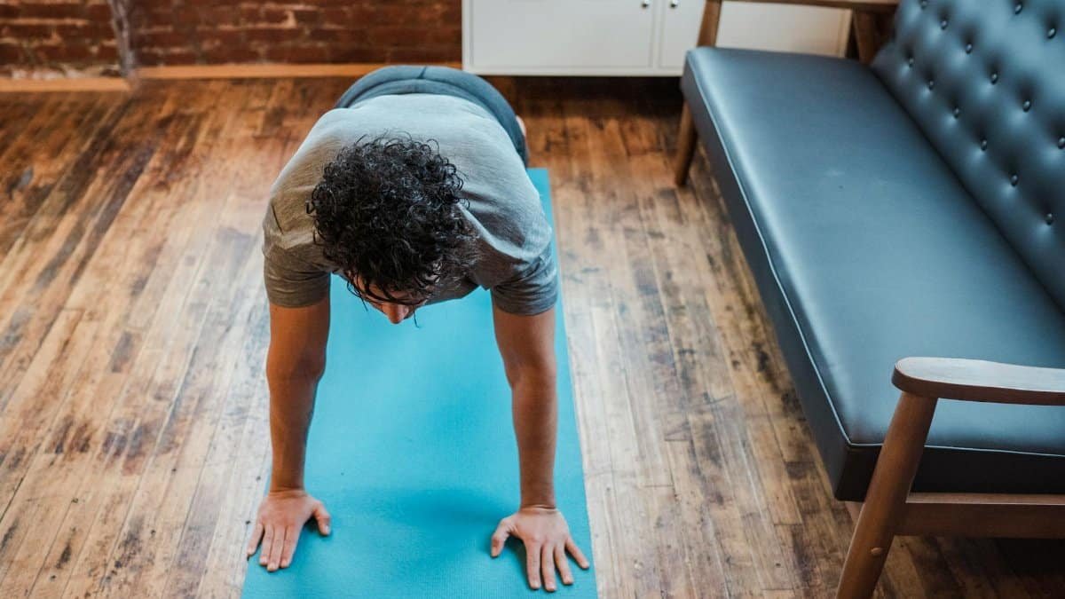High angle of fit male doing balancing yoga posture Phalakasana for strengthening arms on sports mat near sofa