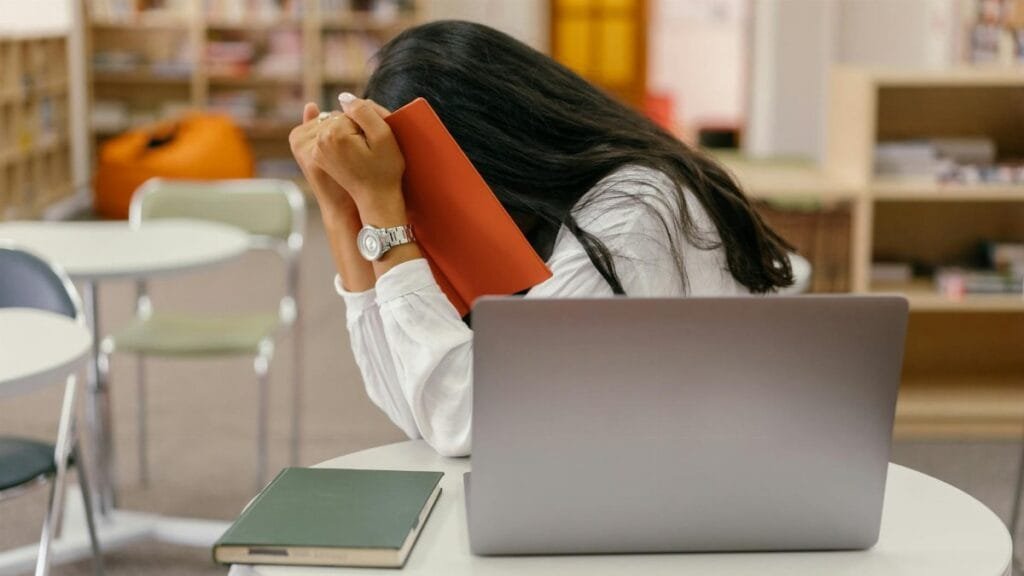 A student hides her face in a book, sitting at a library desk with a laptop, conveying stress.