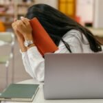 A student hides her face in a book, sitting at a library desk with a laptop, conveying stress.