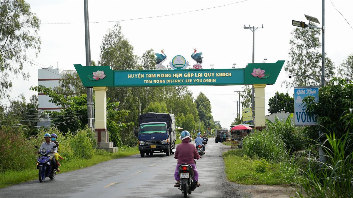 Motorbikes and vehicles travel under Tam Nong District sign on a vibrant rural road.