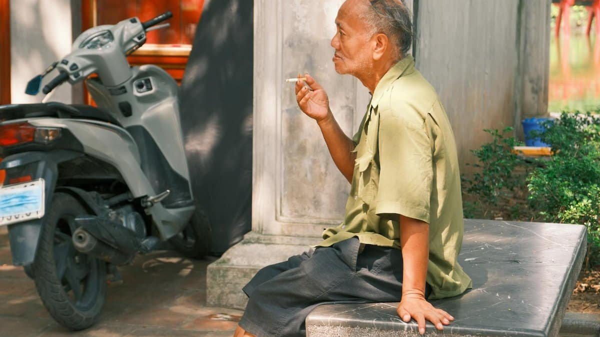 An elderly man smoking while seated on a bench beside a scooter in Hanoi, capturing the essence of daily life.