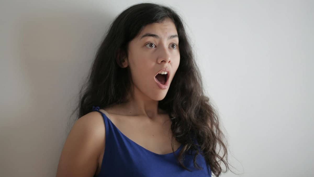 A surprised young woman with long brunette hair and an expressive face in a blue top.