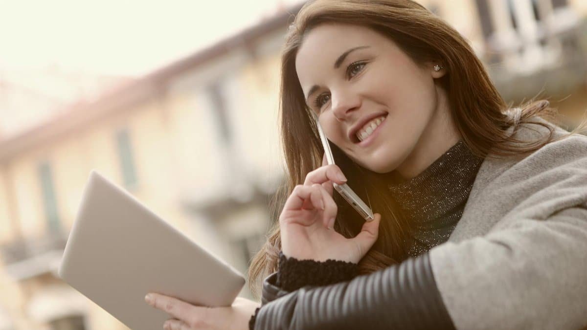 A smiling woman multitasking with a smartphone and tablet outdoors in daylight, showcasing modern technology and lifestyle.