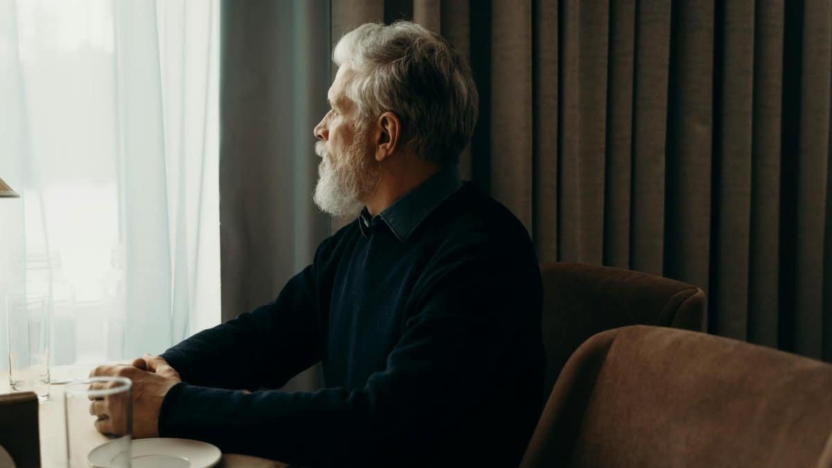 An elderly man with a beard looking outside while sitting by a table indoors.