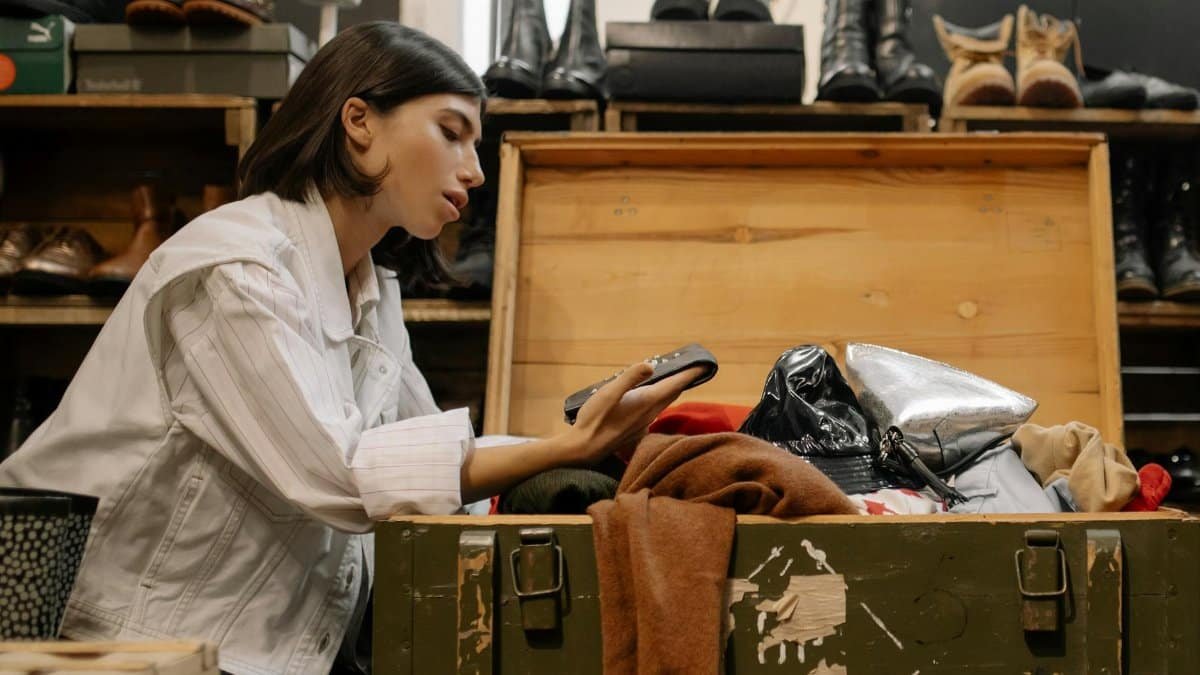 A woman examines clothes from an open chest in a thrift store filled with shoes.