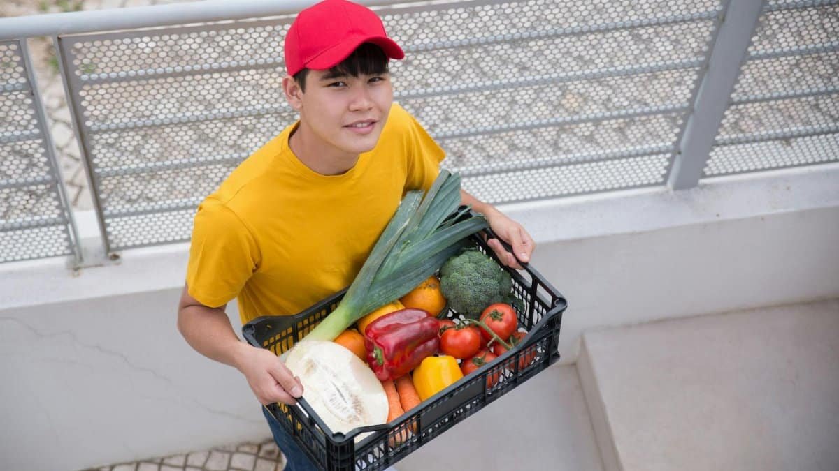 Young man holding a crate of fresh vegetables outdoors, wearing a red cap and yellow shirt.