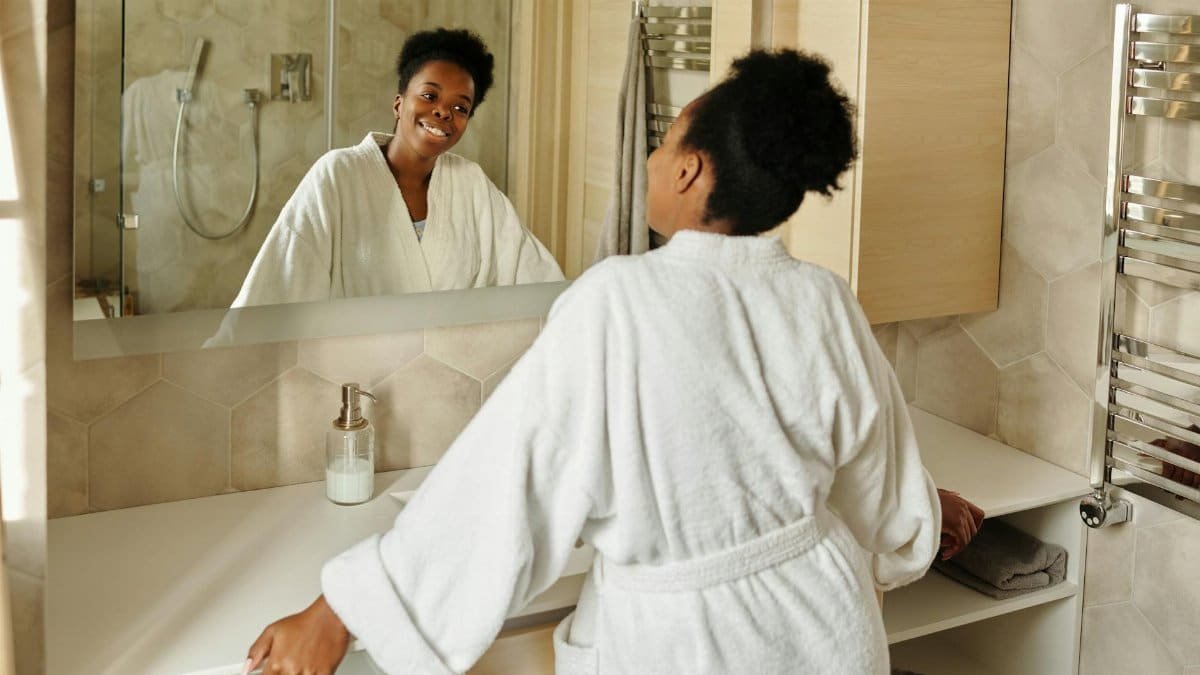 A woman in a bathrobe smiling at her reflection in a modern bathroom mirror.