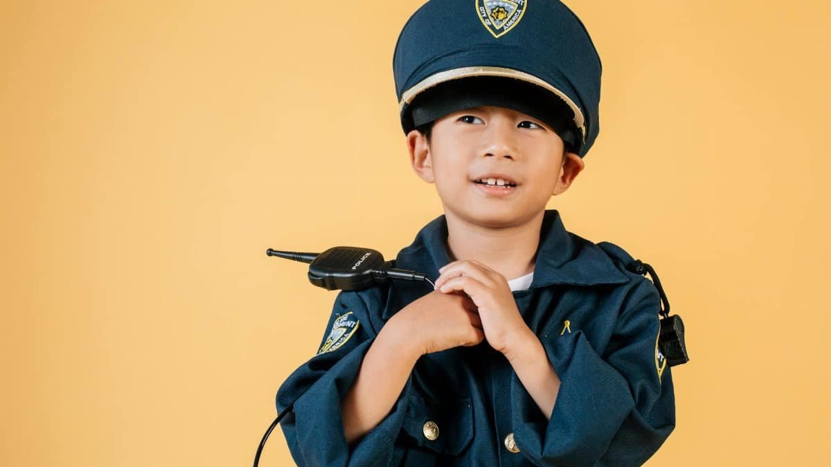 Pleasant Asian boy in police uniform and cap looking away while standing with hands near chest in studio on yellow background