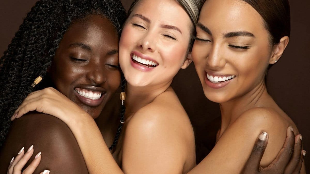 Three diverse women embracing with eyes closed and smiling, celebrating friendship and unity.