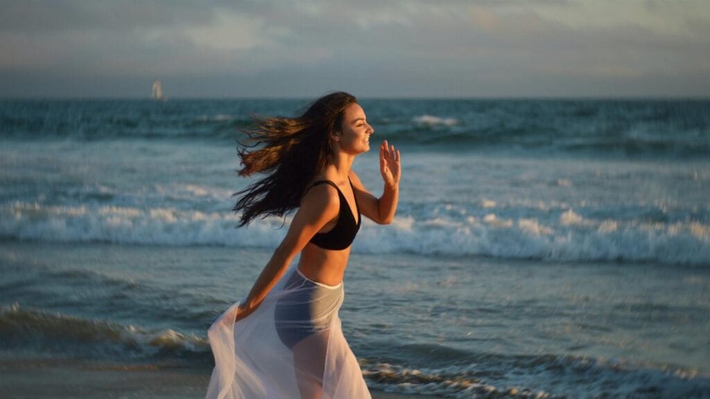 Side view of young happy female tourist with long dark hair in swimwear and pareo smiling and running on beach towards waving ocean during summer holidays