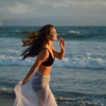 Side view of young happy female tourist with long dark hair in swimwear and pareo smiling and running on beach towards waving ocean during summer holidays