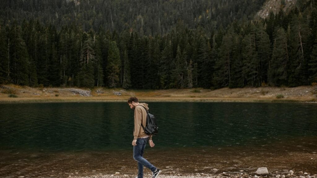 A lone man enjoys a walk by a tranquil mountain lake, surrounded by lush forest scenery.