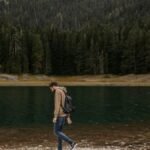 A lone man enjoys a walk by a tranquil mountain lake, surrounded by lush forest scenery.