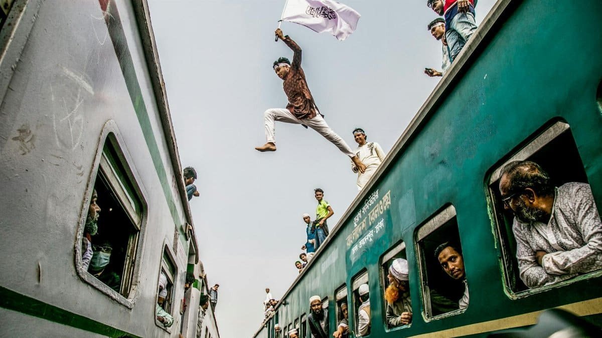 Man leaps between moving trains during daytime amidst a crowded railway scene.