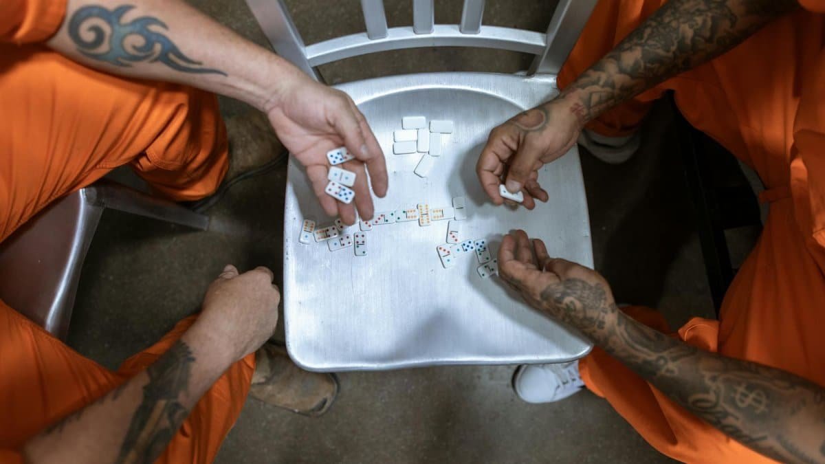 Two tattooed inmates in orange uniforms playing dominoes on a chair in prison.