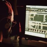 Back view of anonymous bearded freelancer in headphones working on computer while using professional program and listening to music tracks in evening in apartment