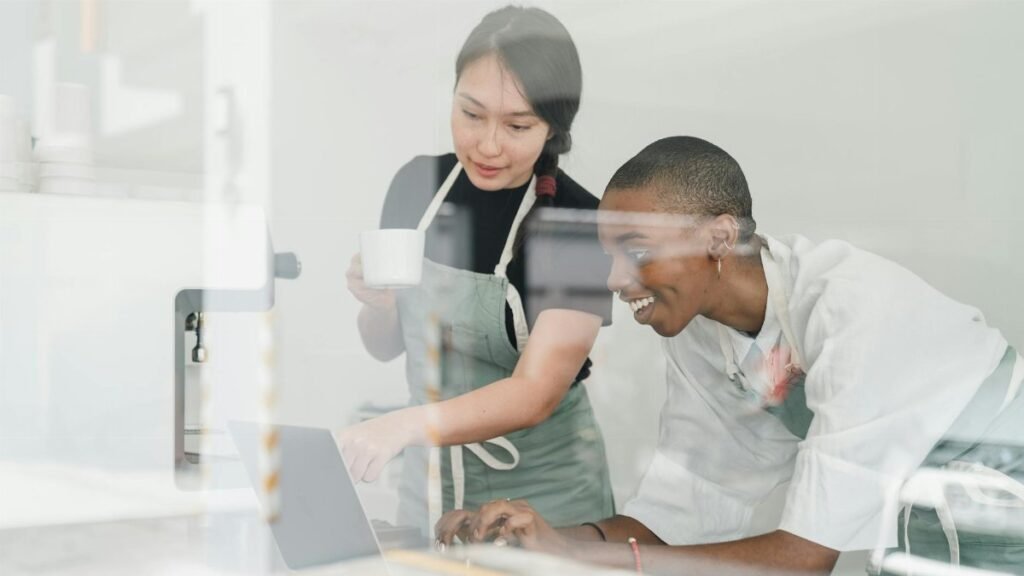 Young inexperienced African American female worker smiling and looking at laptop screen and Asian head barista explaining how special program works both shot through glass
