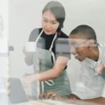 Young inexperienced African American female worker smiling and looking at laptop screen and Asian head barista explaining how special program works both shot through glass