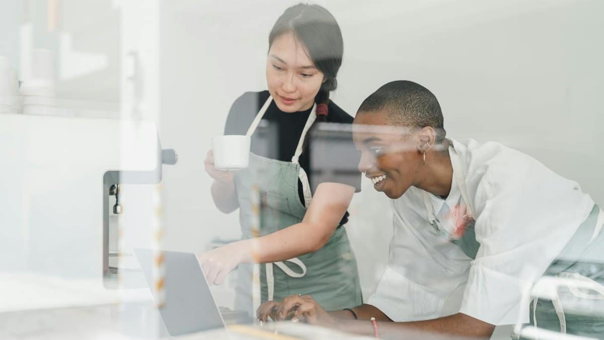 Young inexperienced African American female worker smiling and looking at laptop screen and Asian head barista explaining how special program works both shot through glass