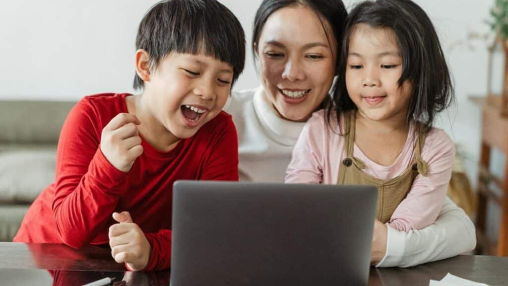 Cheerful little Asian boy and girl watching funny video on laptop with happy smiling mother during weekend