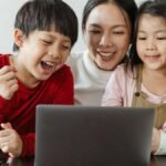 Cheerful little Asian boy and girl watching funny video on laptop with happy smiling mother during weekend