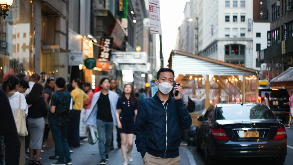 A man wearing a face mask walks down a busy city street, using a phone, surrounded by pedestrians and cars.