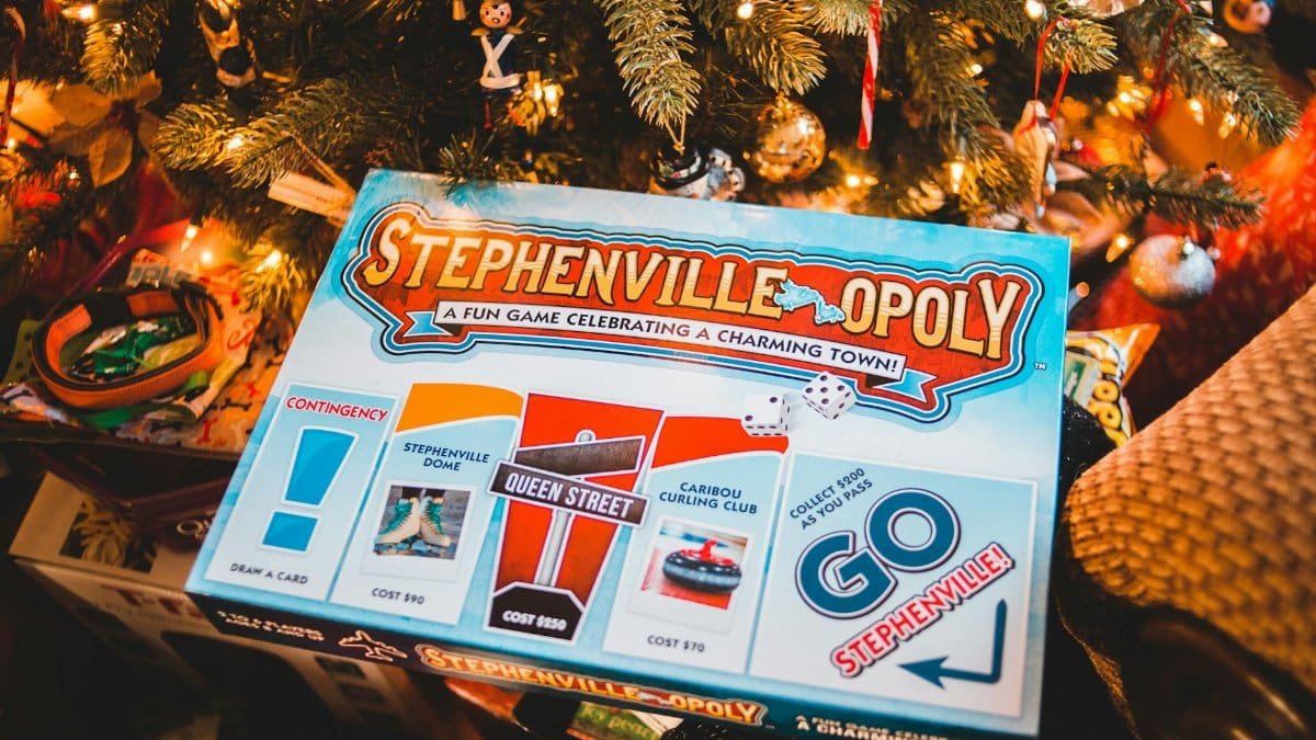 High angle of various gifts and box of creative tabletop game placed near decorated fir tree with glowing garlands during Xmas holidays