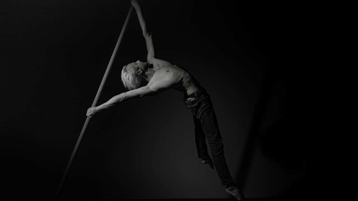 A man performing a dramatic dance move with a pole in a black and white studio setting.