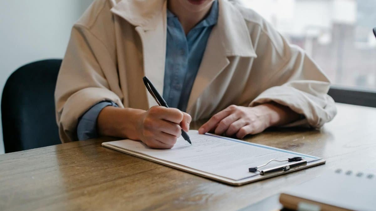 A woman in a formal setting fills out paperwork on a clipboard at an office desk.