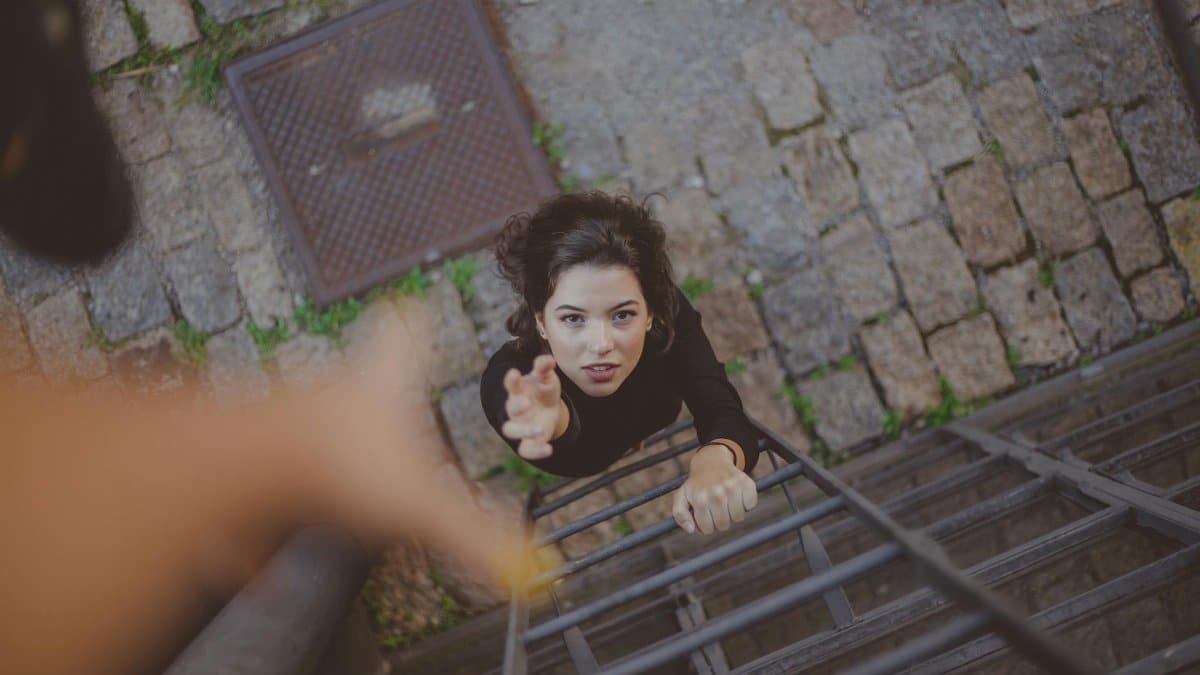 Woman climbs iron ladder outdoors, reaching for help with determination.