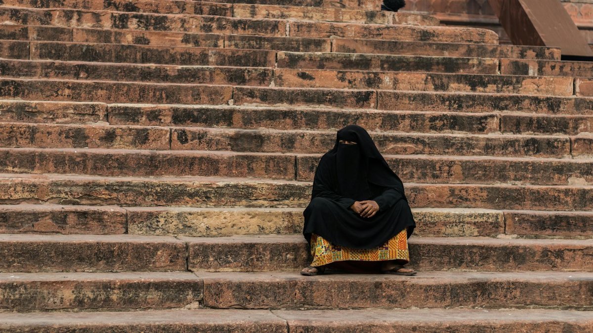 Muslim woman in black abaya sitting on historic stone steps in Delhi, India.