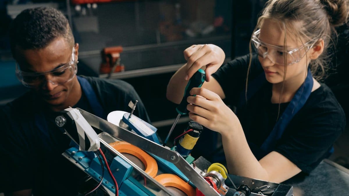 Two students working together on building a robot in an industrial setting, wearing protective goggles.