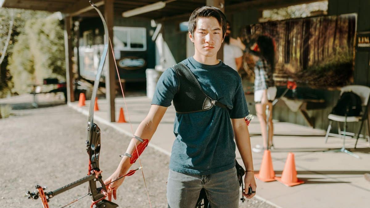 A young adult man practicing archery at an outdoor range on a sunny day.
