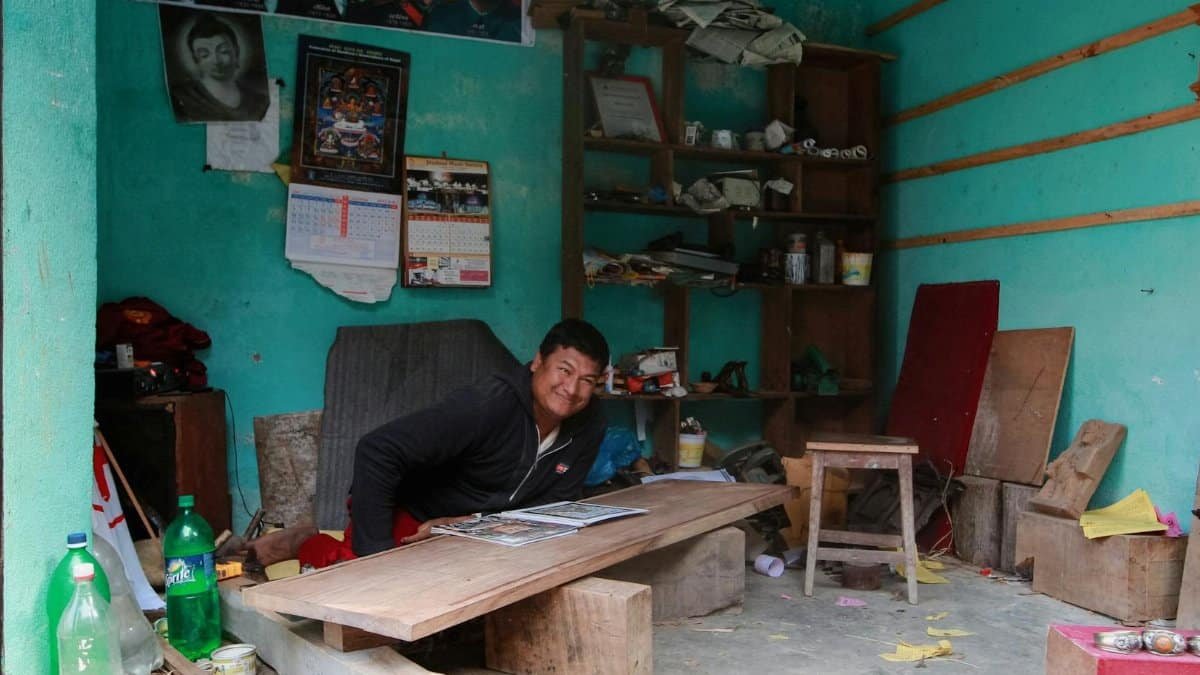 A smiling man sits in a vibrant workshop surrounded by tools and vibrant posters, exuding creativity and industry.