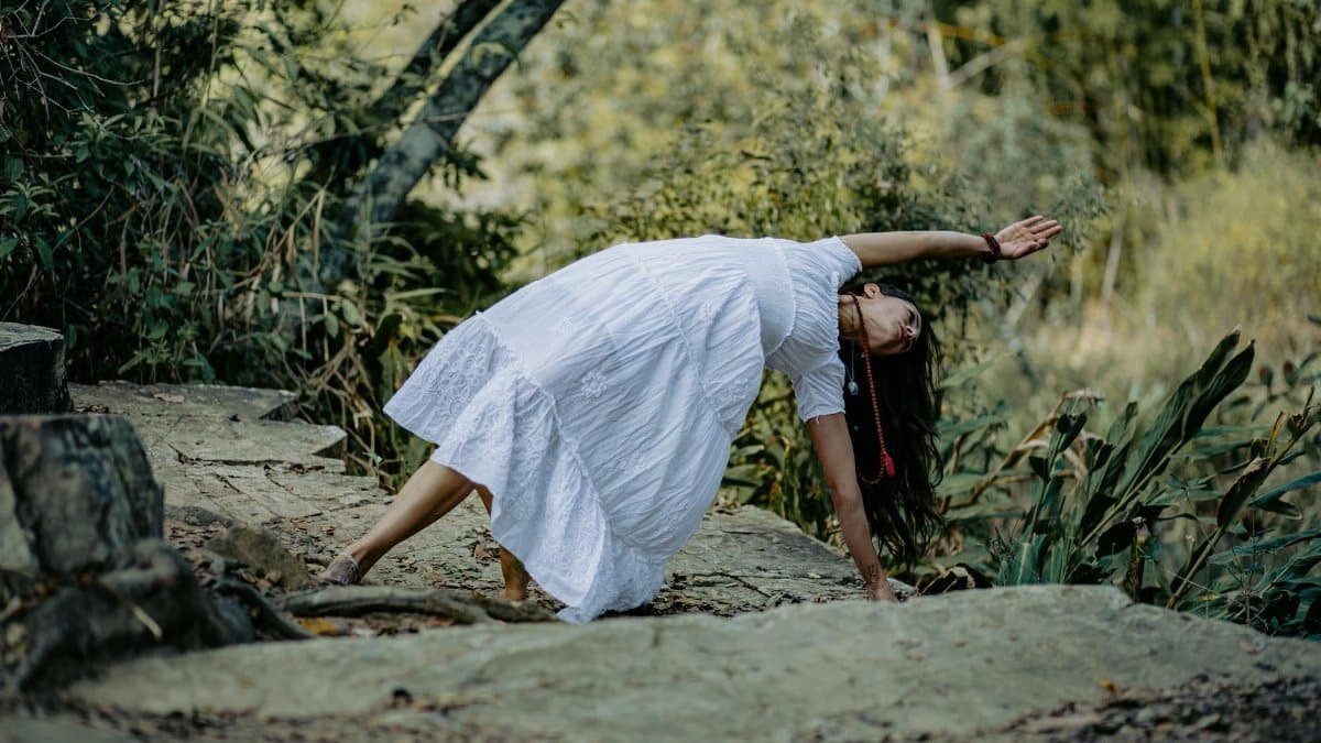 Woman in white dress practicing yoga on stone path in a lush forest setting.
