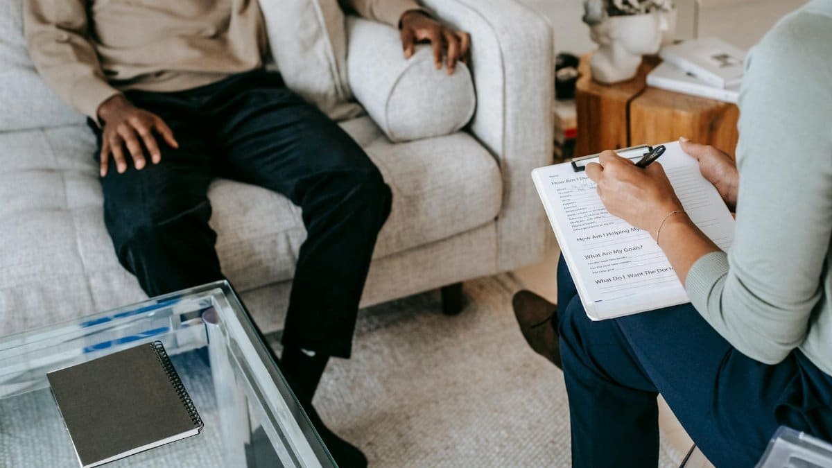 From above crop African American male in casual clothes sitting on comfy couch and answering professional female consultant questions in modern psychotherapy office