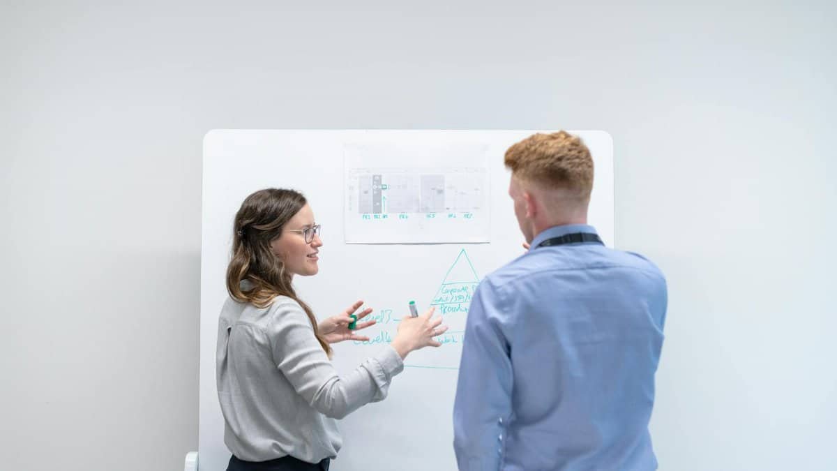 Two professionals discussing project plans at whiteboard in office setting.
