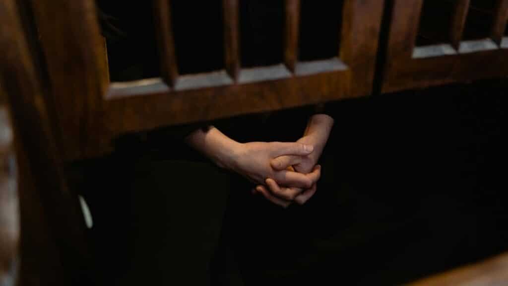 Hands clasped in prayer inside a wooden confessional, symbolizing faith and introspection.