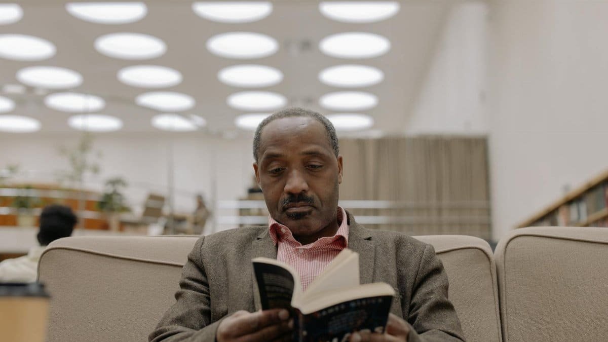 An African American man seated on a couch reading a book in a modern library setting with unique ceiling lights.