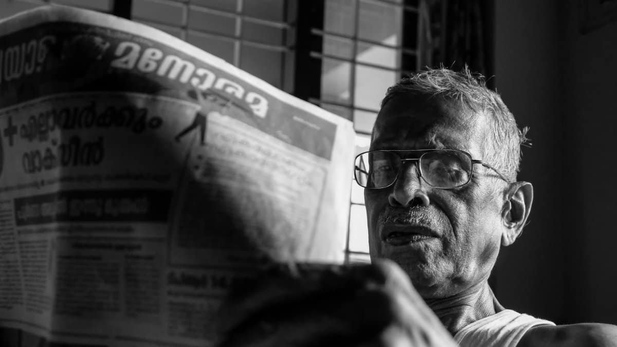 Black and white photo of an elderly man with glasses reading a newspaper in Nilambur, India.