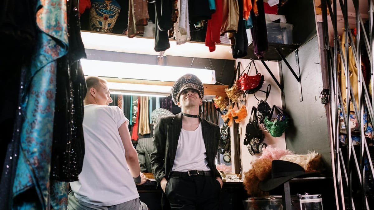Two performers prepare in a theatrical dressing room, featuring costumes and vibrant attire.