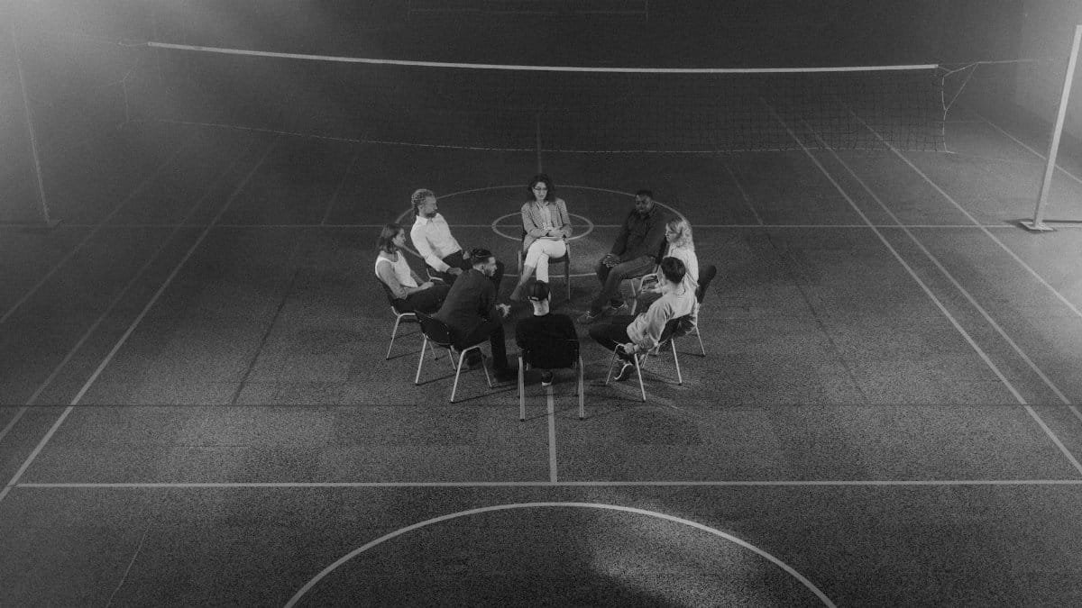 A therapy group sitting in a circle in a dimly lit sports hall.