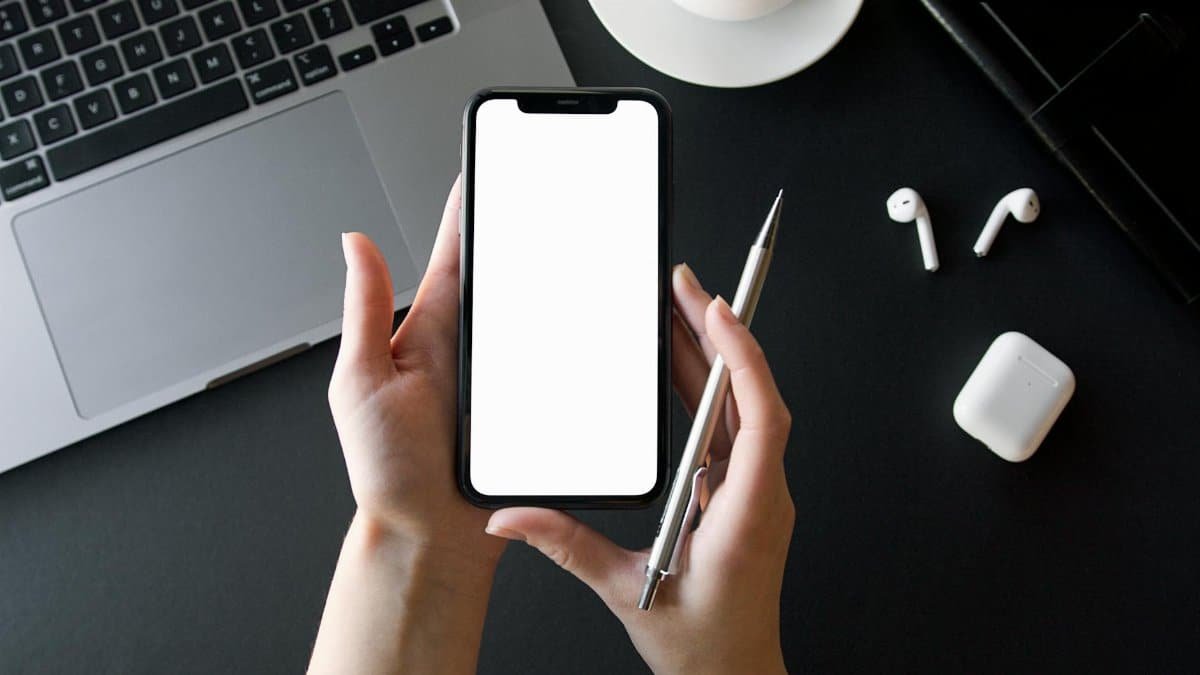 Close up of hands holding a smartphone over a desk with laptop and earbuds.