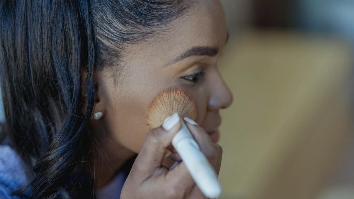 Side view of a young woman applying makeup indoors with a brush.