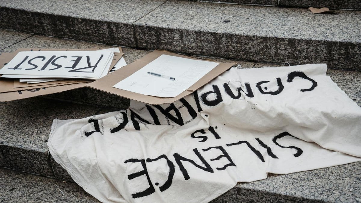 A close-up of protest signs reading 'RACIST' and 'Silence is Complacency' displayed on stone steps.