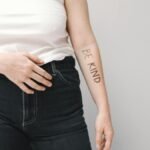Person in jeans and white tank top with 'Be Kind' text on arm, promoting kindness.